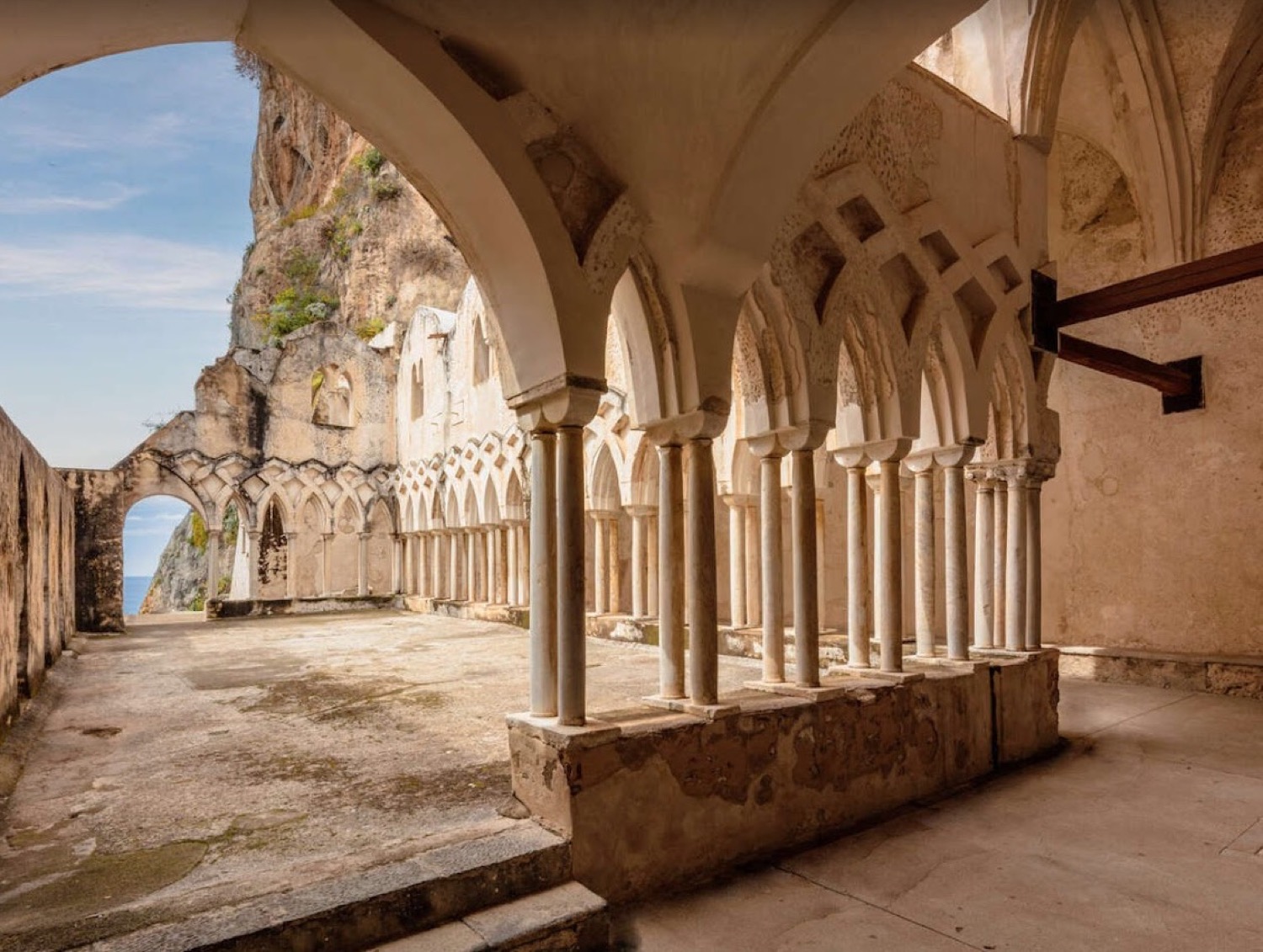 The white Gothic arches of the 13th-century cloister at the Anantara Convento di Amalfi, with the cliffs and sea beyond