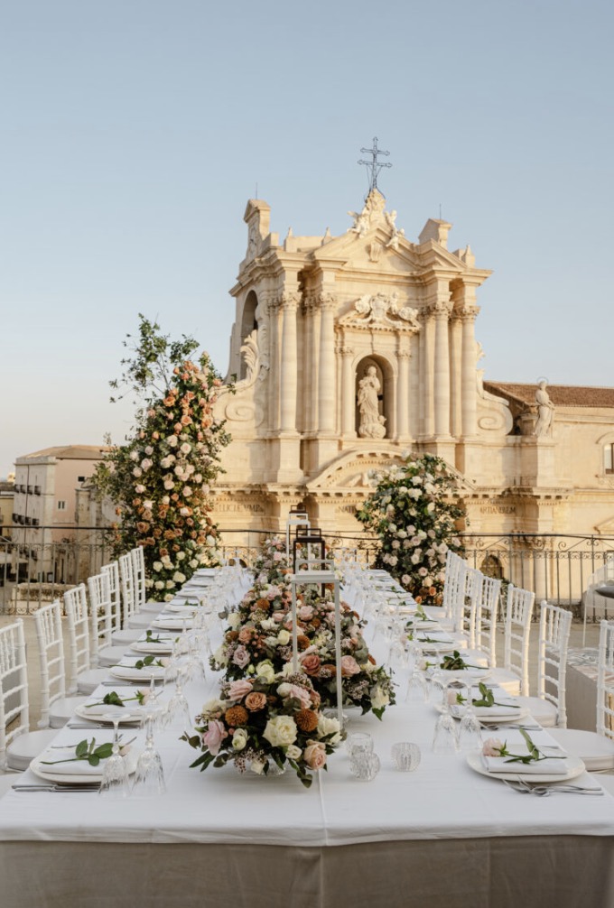 Long terrace table with white linens and rose centerpieces at Terraza Killiche in Sicily, with the historic church facade behind
