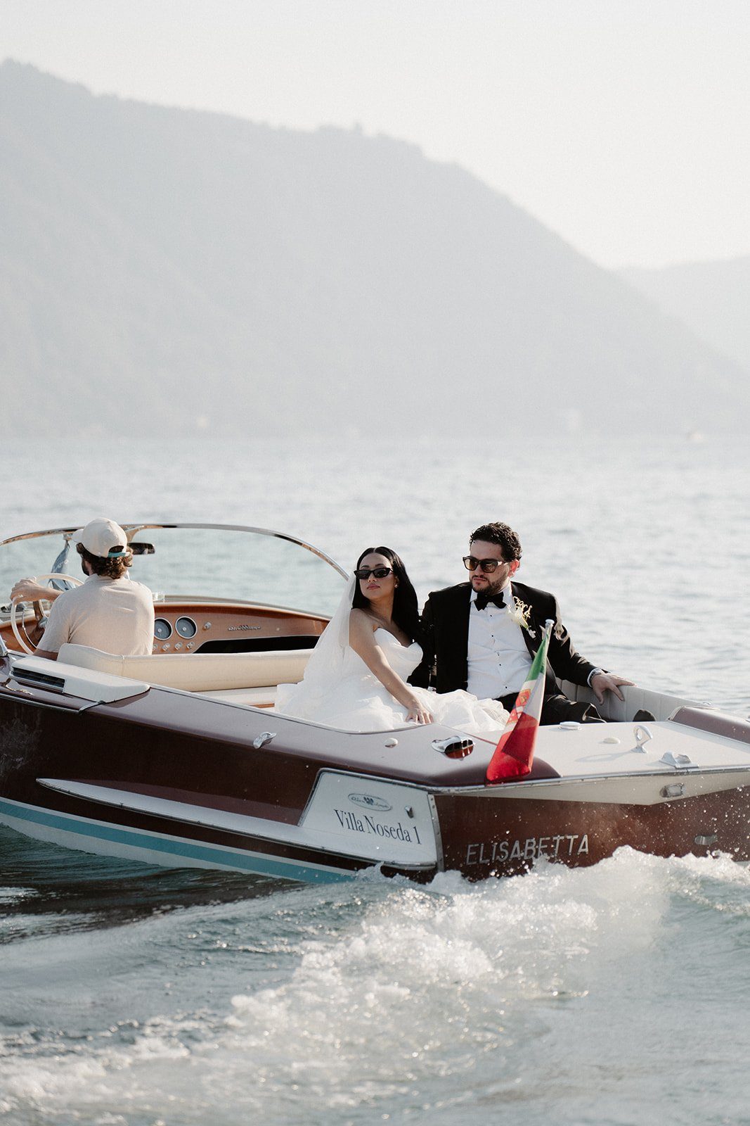 A close view of a couple on a wooden boat, with a bride’s bouquet and the lake behind
