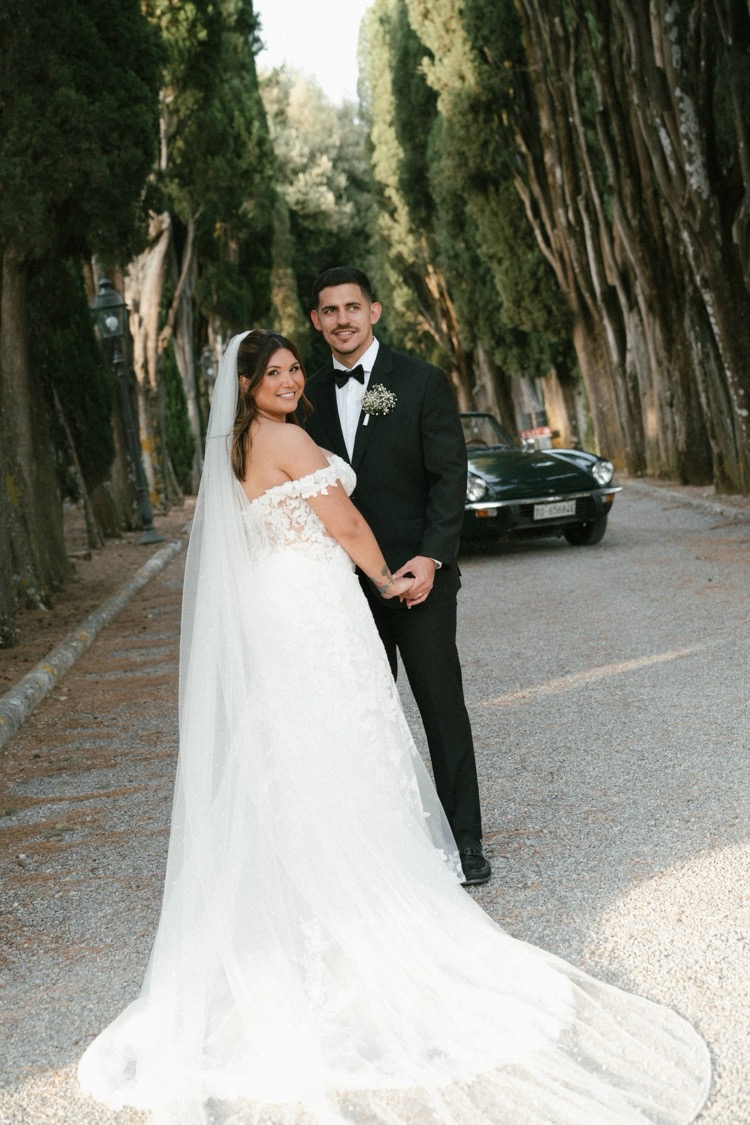 A bride and groom posing on the cypress-lined drive at Villa Poggiano, Tuscany, with a vintage green convertible behind them