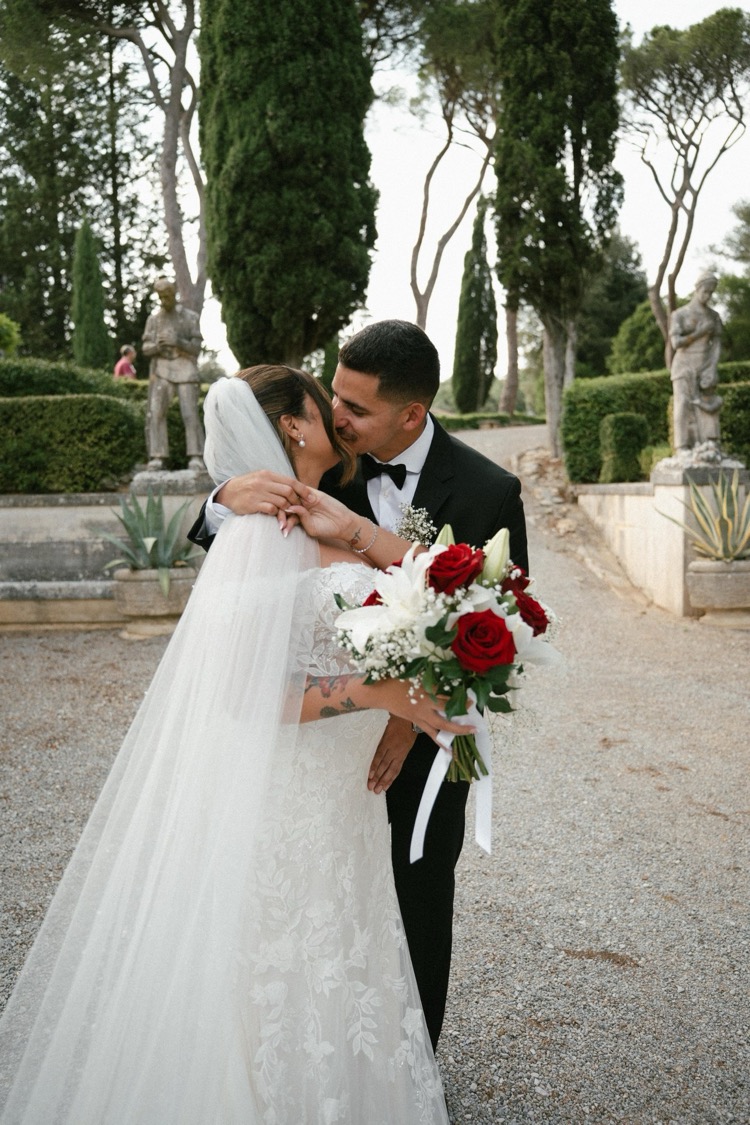 A newlywed couple embracing with a red and white bouquet among the statues and hedges of Villa Poggiano, Tuscany