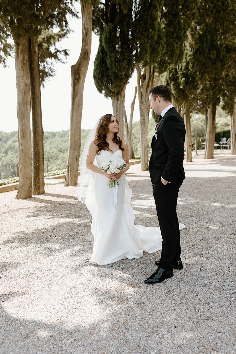 A bride and groom standing together on a tree-lined garden path at Villa Poggiano, Tuscany, with the Tuscan hills beyond