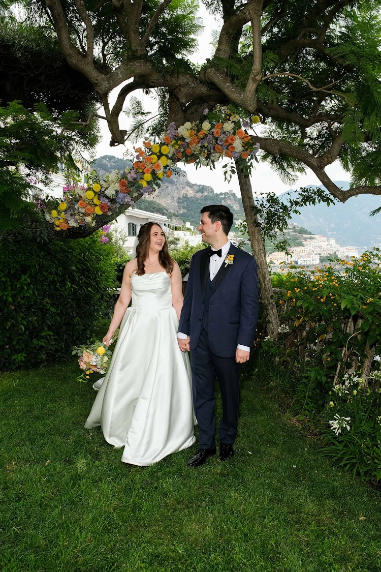 Bride and groom laughing together under a flower arch in the Hotel Santa Caterina garden