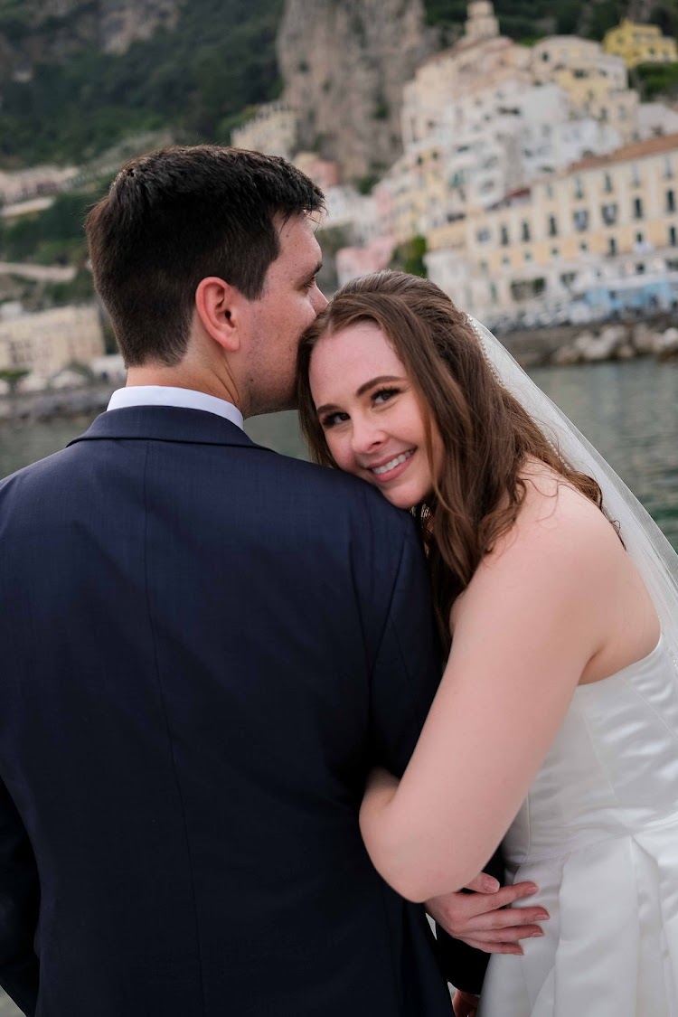 Bride smiling on her groom's shoulder with the town of Amalfi behind, Amalfi Coast